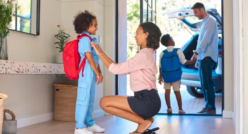 Photo of woman in hallway adjusting backpack of child, background shows man holding hand of child.