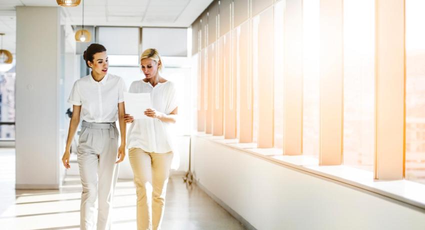 Two women walking by windows in office setting.