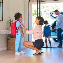 Photo of woman in hallway adjusting backpack of child, background shows man holding hand of child.
