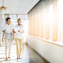 Two women walking by windows in office setting.
