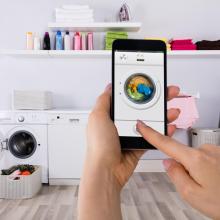 Photo showing hands of person holding smart phone. Smart phone shows photo of a washing machine. Background is laundry room.